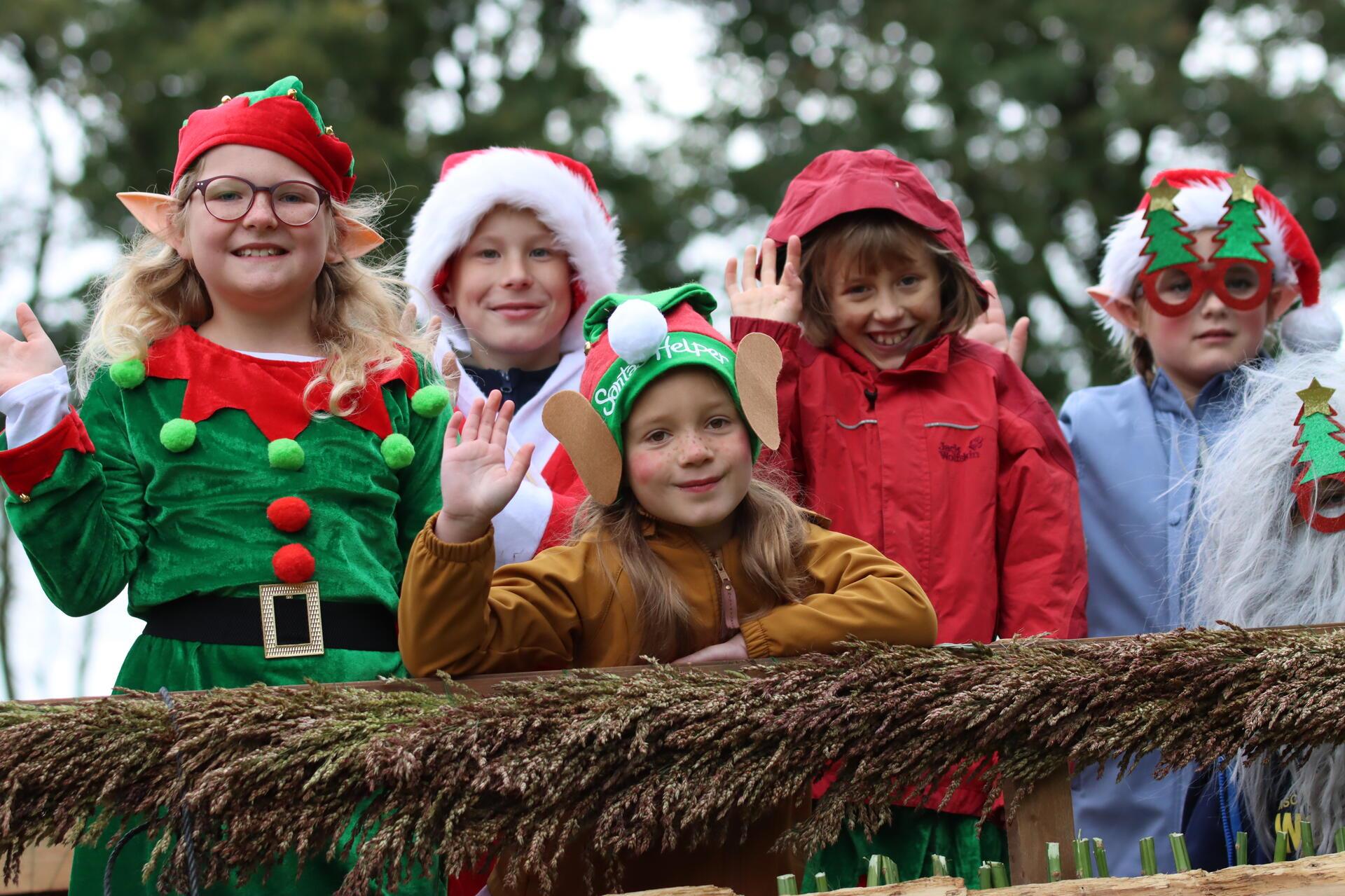 Fünf Kinder in Weihnachtselfen-Kostümen winken von einem Wagen.