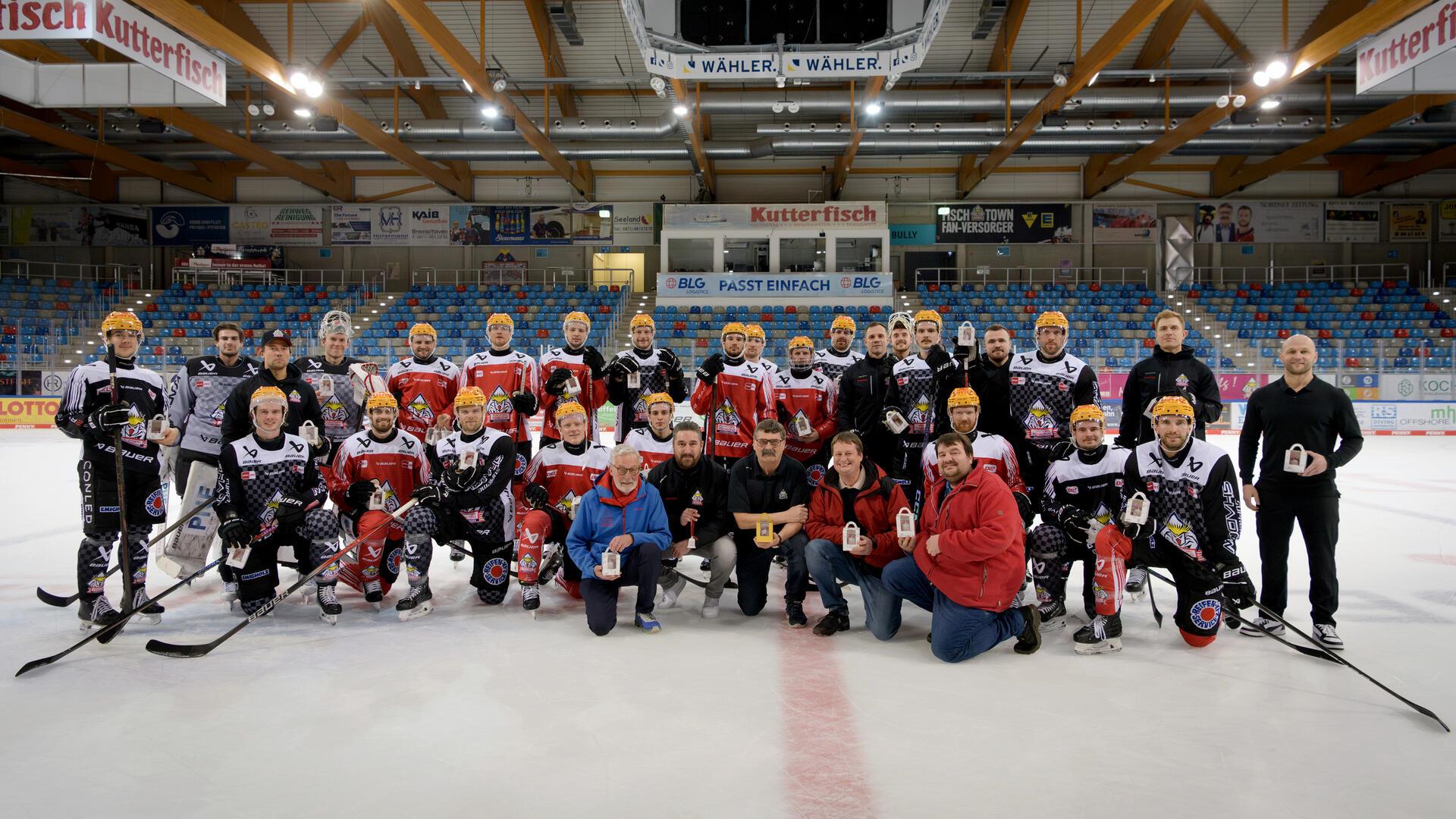 Eine große Gruppe von Eishockeyspielern in roten und schwarzen Trikots sowie Trainer und weitere Personen posieren gemeinsam auf dem Eis einer Eishalle.