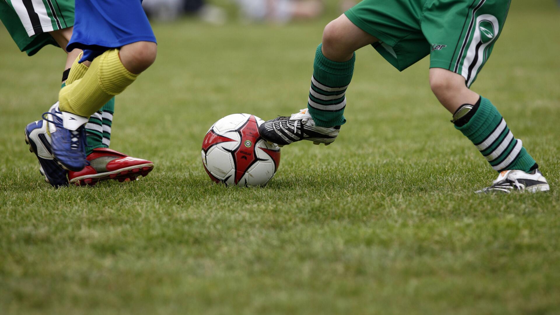 Zwei Kinderbeine in Fußballstutzen und -schuhen mit einem Ball zwischen sich.