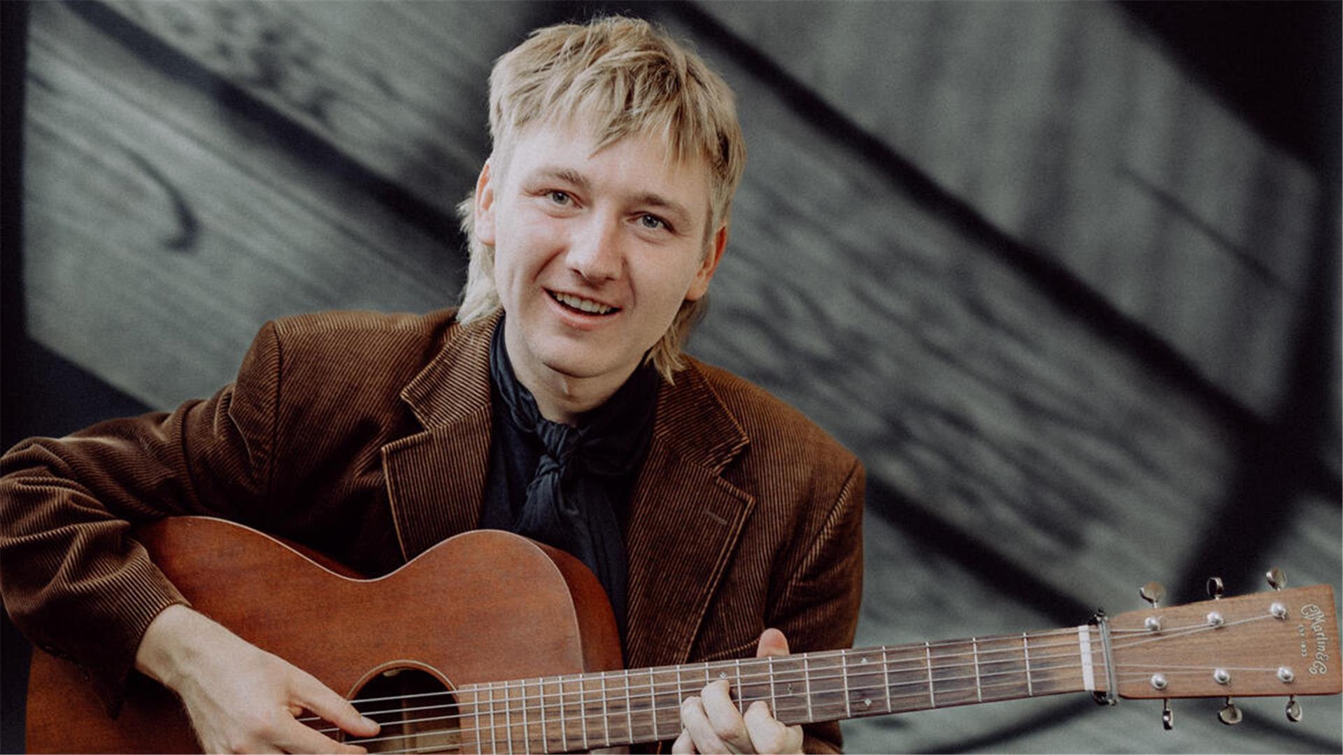 Ein junger Mann lächelt in die Kamera und zupft an seiner Gitarre.