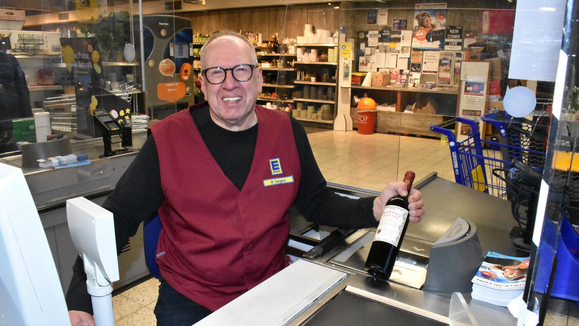 Ein Mann mit einer Brille sitzt an einer Kasse im Supermarkt und hält eine Flasche in der linken Hand.