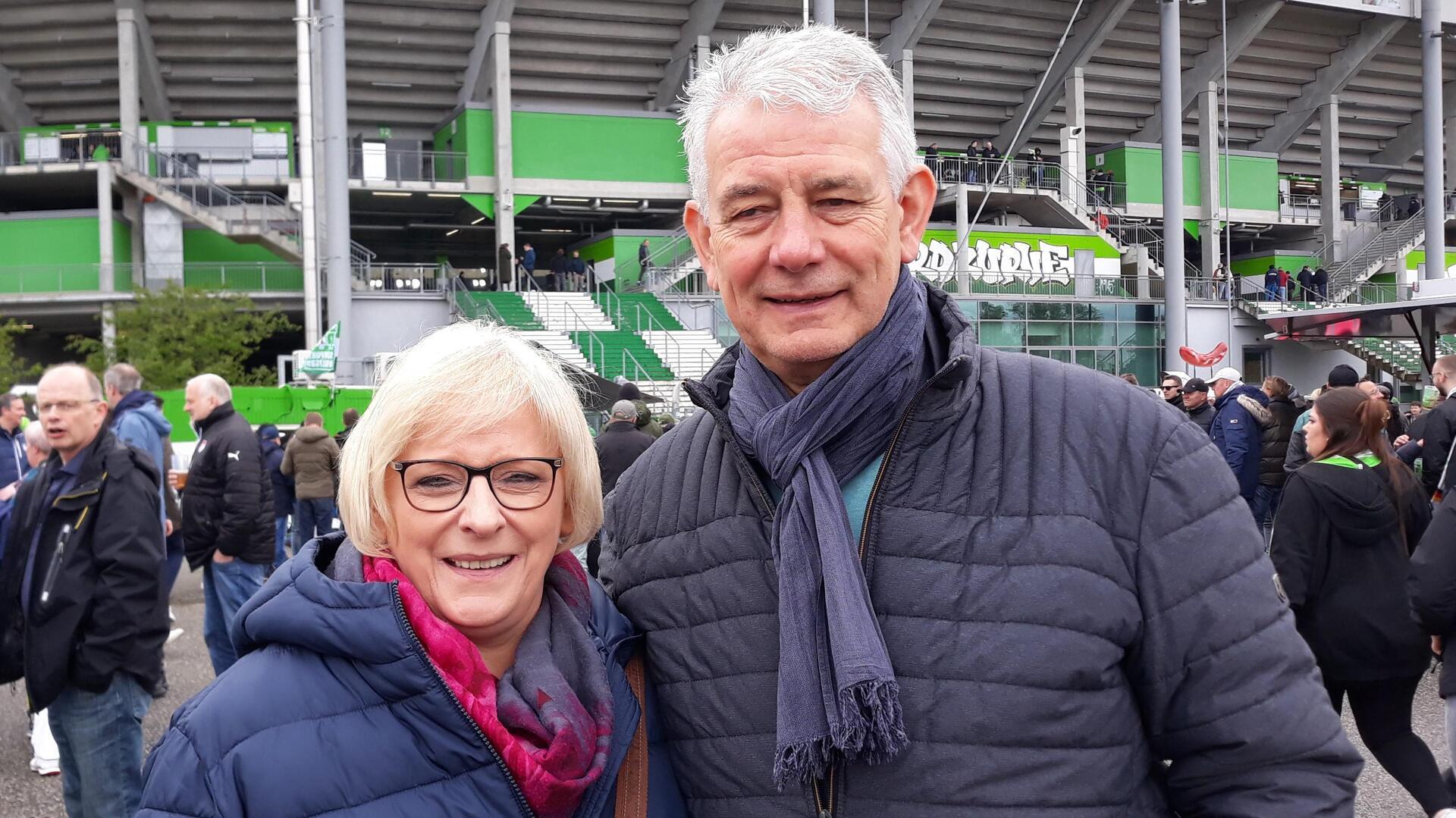 Ein älteres Ehepaar in blauen Daunenjacken steht vor einem Fußballstadion.