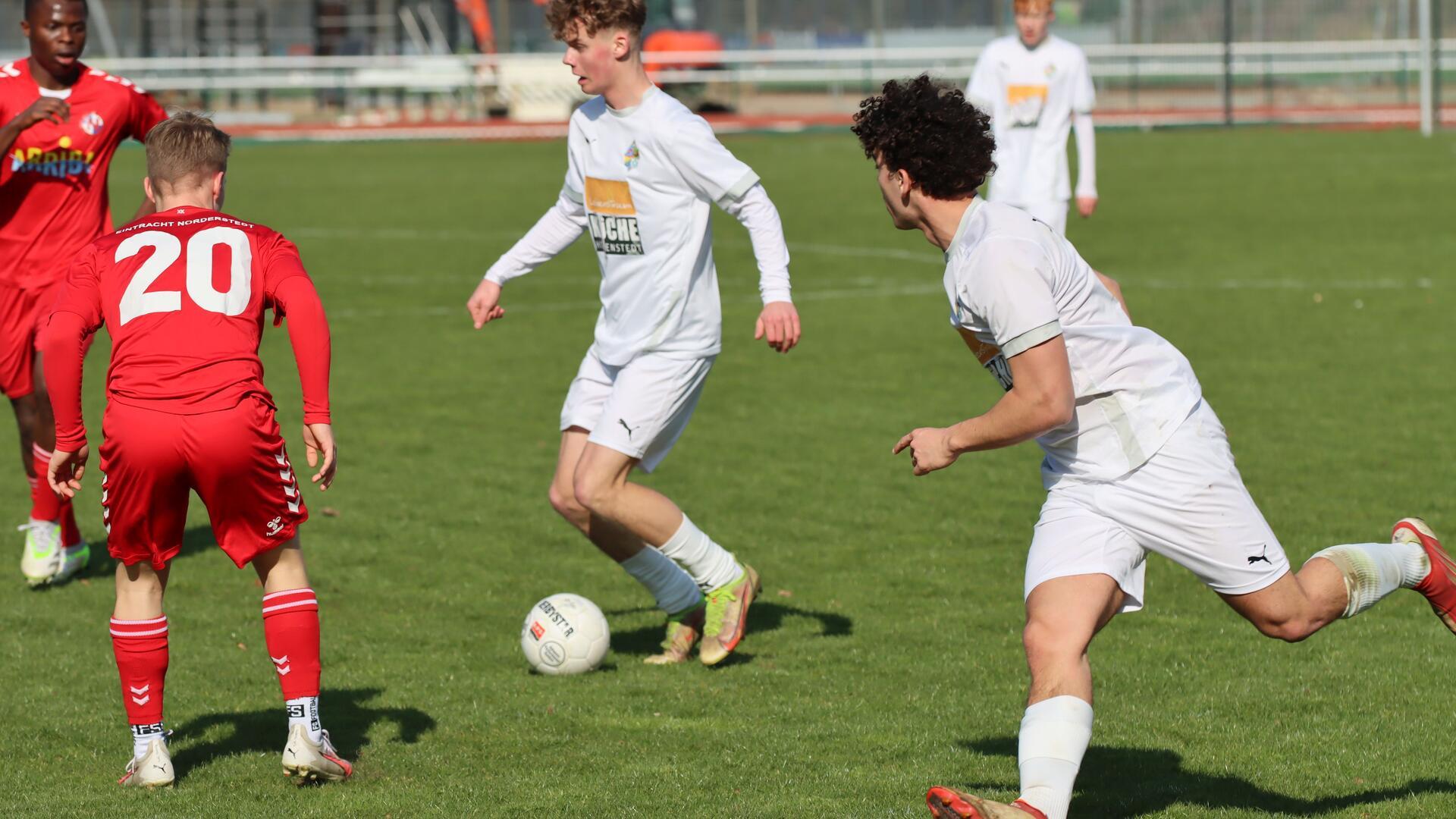 Zwei junge Männer in weißen Trikots laufen hinter einem Ball her, zwei junge Männer in roten Trikots stellen sich in den Weg.