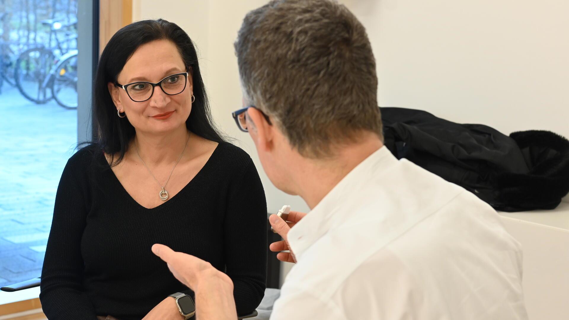 Eine Frau mit schwarzen Haaren und Brille sitzt einem Arzt gegenüber.