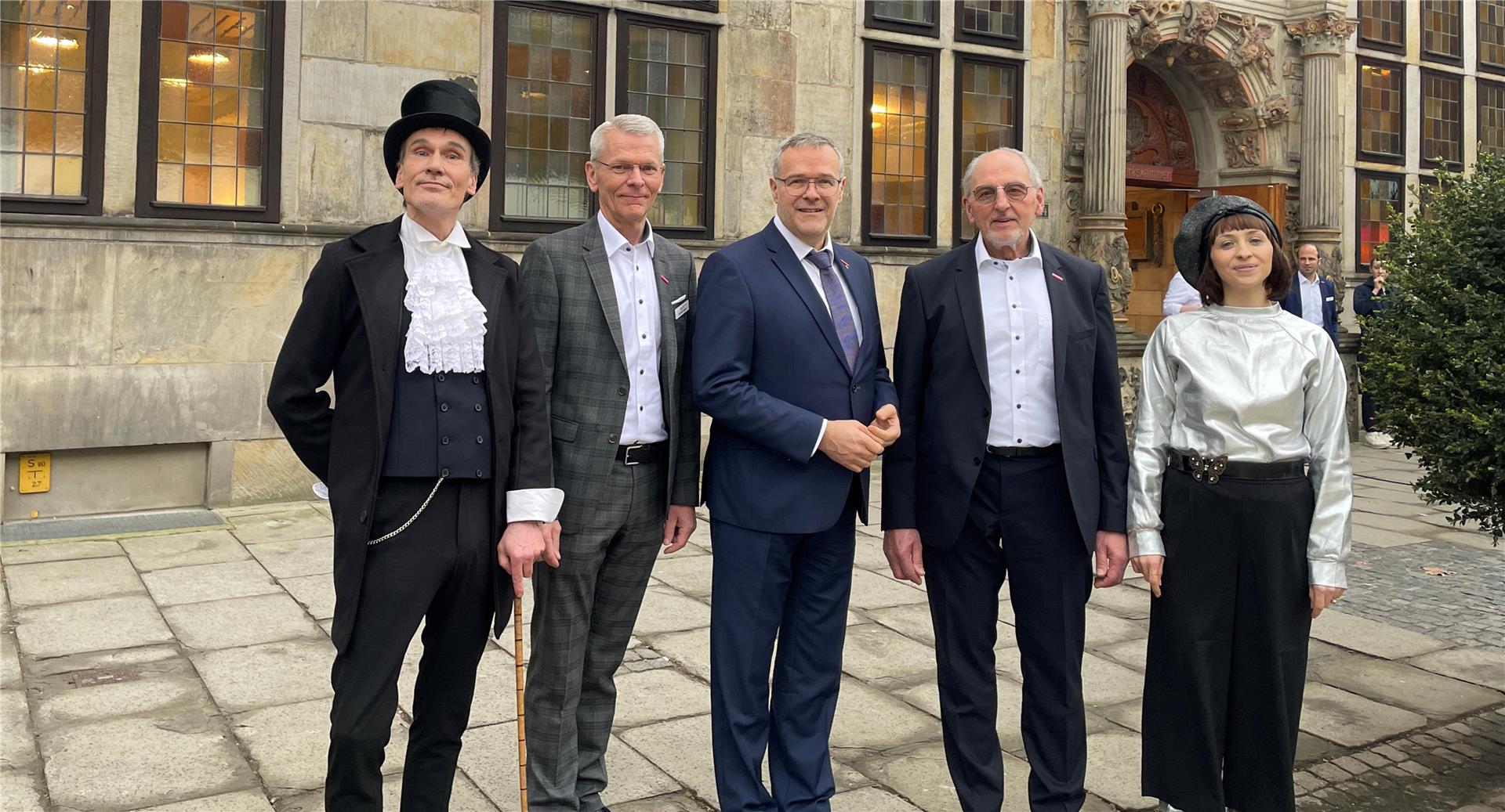 Fünf Menschen, drei in Anzügen, und zwei in Kostümen, stehen vor dem Gewerbehaus der Handwerkskammer Bremen am Ansgarikirchhof.