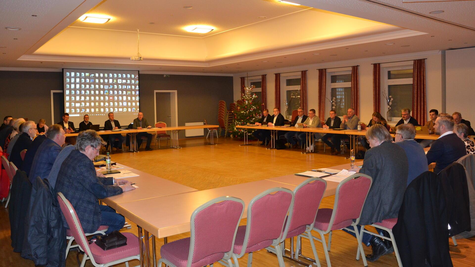 Rund 30 Menschen sitzen im großen Rechteck an Tischen in einem großen Saal.