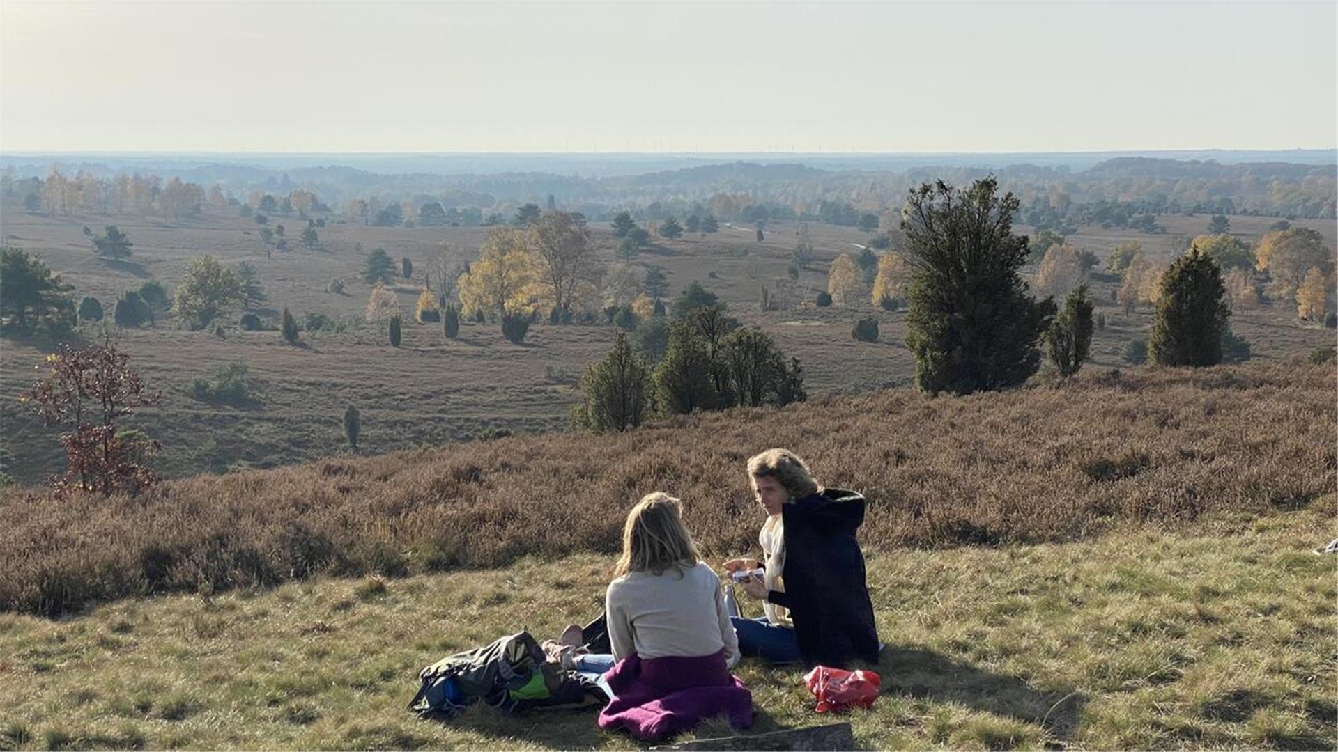 Zwei Personen sitzen auf einer Wiese inmitten einer offenen Heidelandschaft. Vor ihnen liegt eine weite Landschaft mit Bäumen, Sträuchern und sanften Hügeln, die in herbstlichen Farben leuchtet. Der Himmel ist leicht bewölkt.