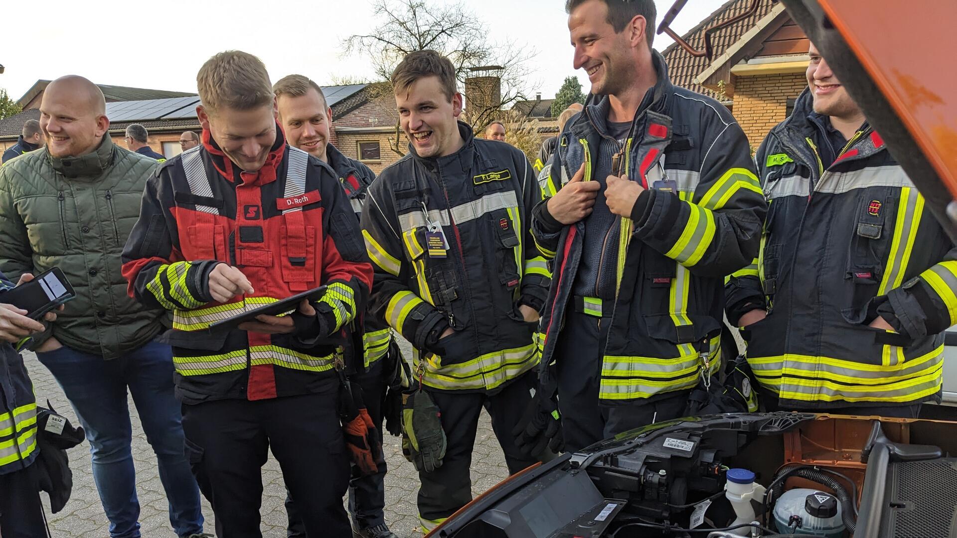 Eine Gruppe von Feuerwehrleuten in Einsatzkleidung steht zusammen und schaut lächelnd auf ein Tablet, das von einem Kollegen in roter Weste gehalten wird. Vor ihnen steht ein geöffnetes Auto mit sichtbarem Motorraum.